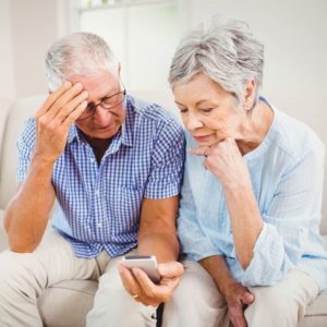 Sad senior couple sitting on sofa and looking at mobile phone in living room