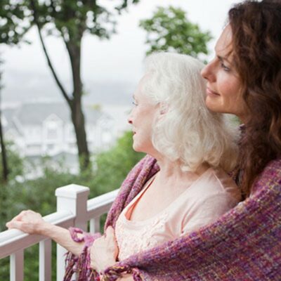 An adult daughter embracing her elderly mother while both are staring off into the distance from a balcony.