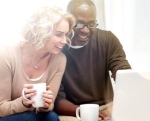 Pareja de esposos interracial con tazas de té; el hombre está señalando a algo en una computadora y sonriendo