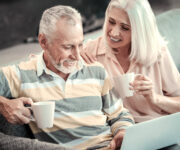 An Elderly Couple sitting on a couch, looking at a laptop while having coffee.