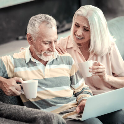 An Elderly Couple sitting on a couch, looking at a laptop while having coffee.